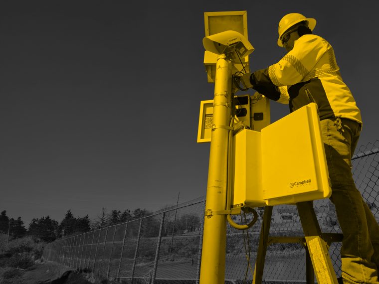A technician servicing a roadside weather station enclosure from a ladder.